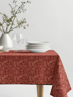 Mantel de hule Ophelia con flores y hojas ornamentales en terracota y rojo oscuro, sobre una mesa de comedor