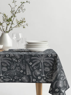 Mantel de hule Tigre pashu con fondo gris oscuro y dibujos en blanco de tigre, palmeras y helechos, sobre una mesa de comedor