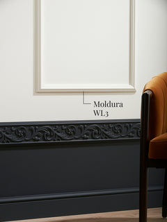 Moldura muro WL3 enmarcando un paño blanco sobre un zócalo oscuro con ornamento, junto a un sillón